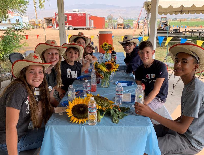A group of teens, all wearing straw cowboy hats, sit at a table outside.
